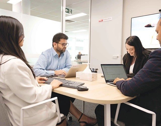 Four professionals in a modern office conference area, working on laptops around a round table and collaborating.